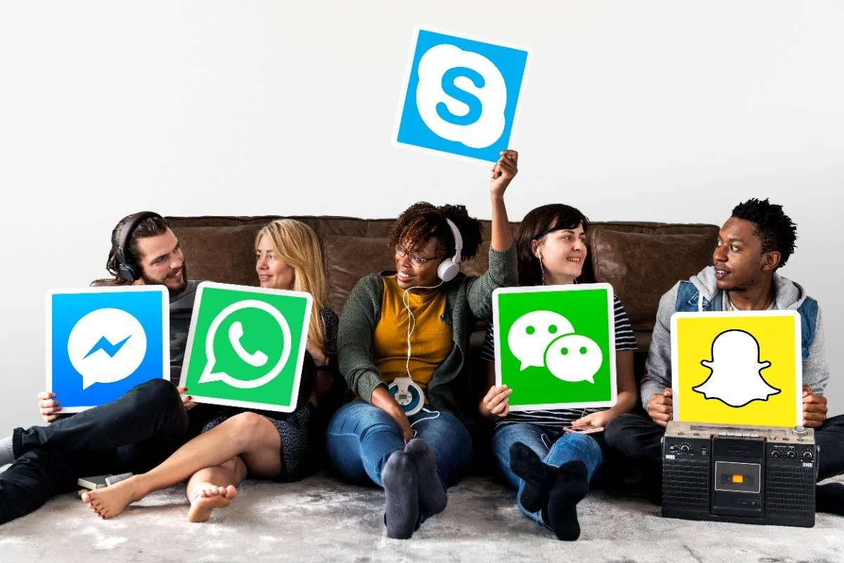 "A group of friends sitting on a couch, each holding signs with logos of popular social media and messaging apps, including Facebook Messenger, WhatsApp, Skype, WeChat, and Snapchat."