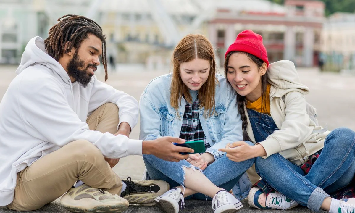 Teenagers sitting outdoors sharing SMS short forms on a smartphone.